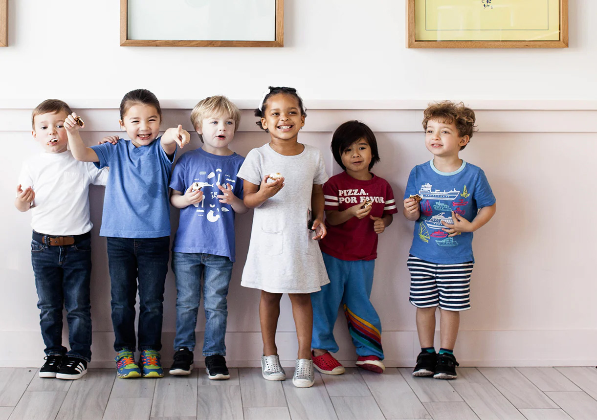 Six children ages 4-6 of various ethnicities standing in a row, eating, smiling, and gesturing