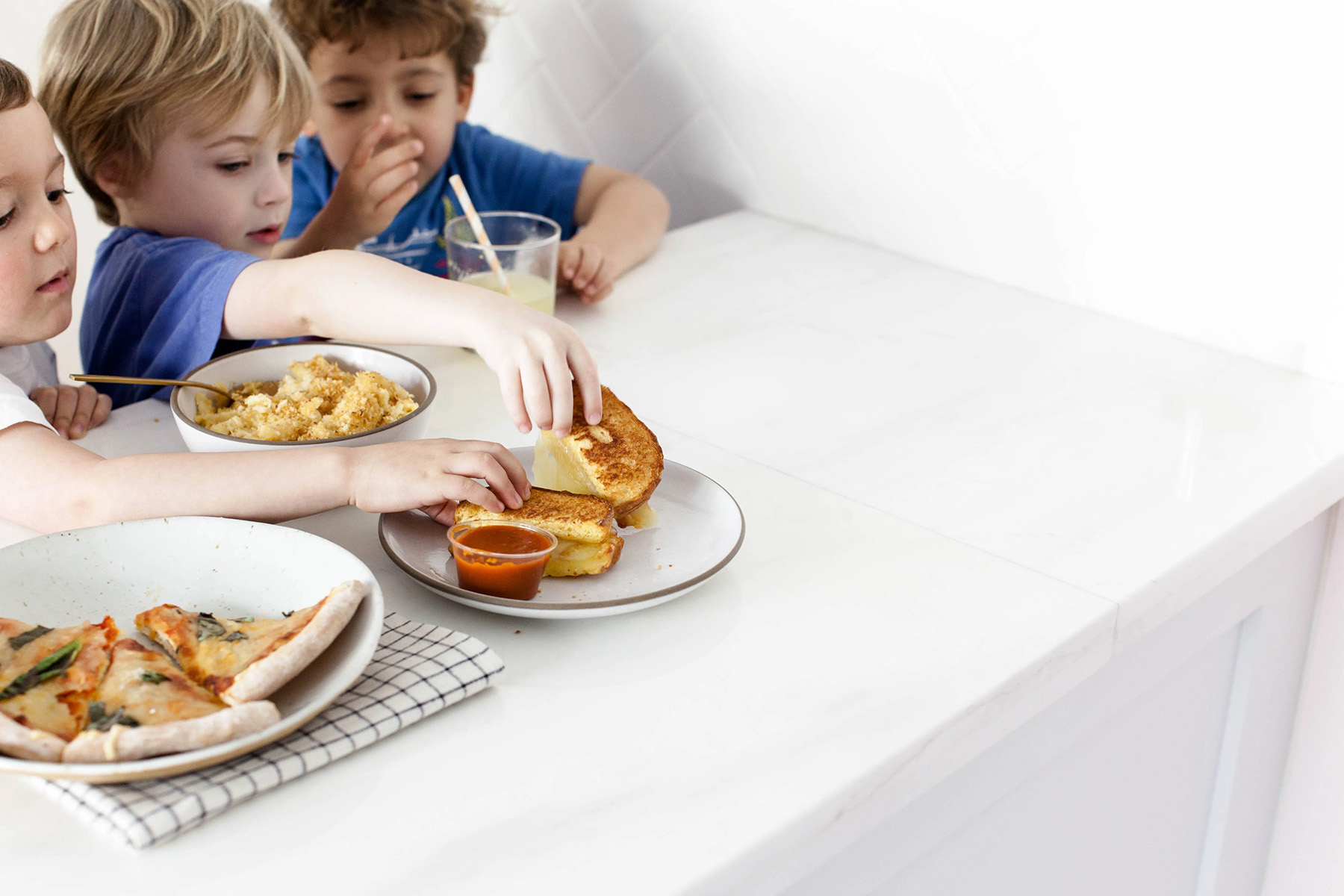 Three kids enjoying a snack of pizza, pasta, and mac and cheese. They're reaching for more food, as they sit up next to a white countertop.